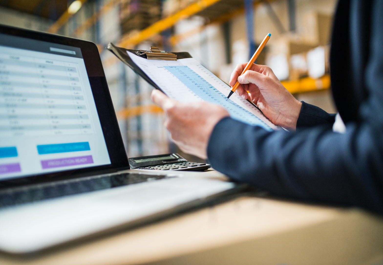 Employee analyzing information on his laptop and clipboard in the warehouse