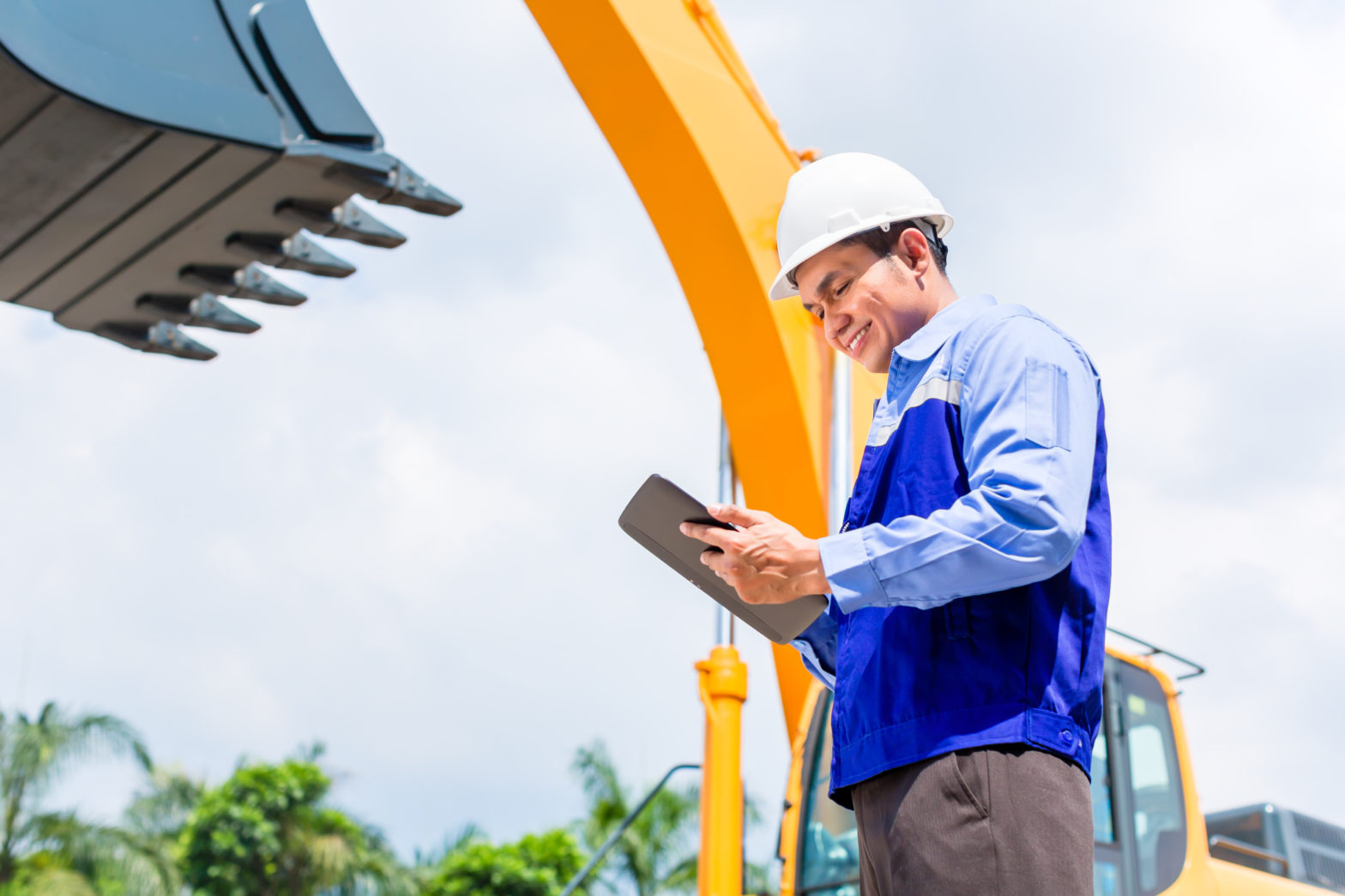 Male employee in hardhat holding and looking at data on a tablet in front of heavy machinery