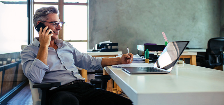Mature male business man sitting at his desk in front of his laptop, smiling while on the phone
