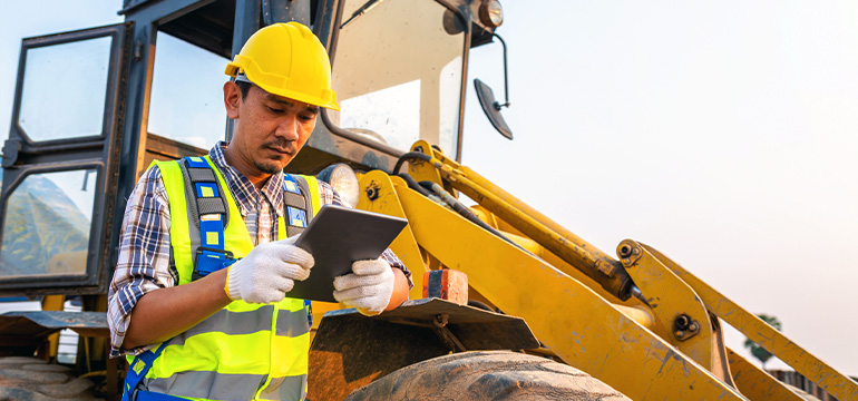 Male in full safety gear outside reviewing data on a tablet next to a piece of heavy equipment