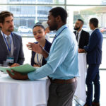 Group of business professionals at a conference, focusing on 3 having a conversation while standing by a table