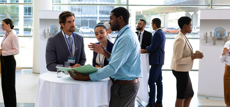 Group of business professionals at a conference, focusing on 3 having a conversation while standing by a table