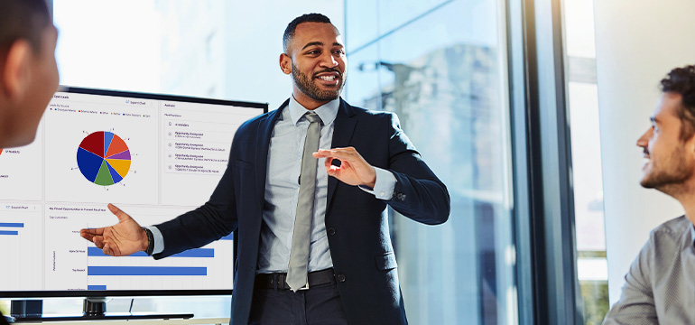 Male business professional speaking to other coworkers and providing a presentation of Microsoft Dynamics 365 sales data on a monitor