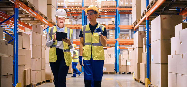 Male and female warehouse workers in safety gear having a conversation and walking holding a tablet