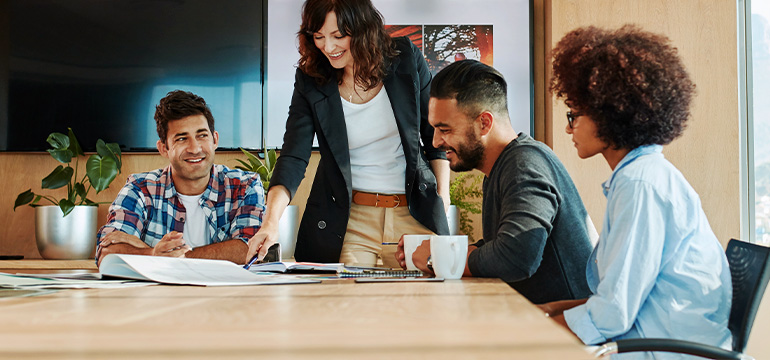 Female coworker standing and pointing at papers amongst 3 other coworkers who are sitting at a large desk